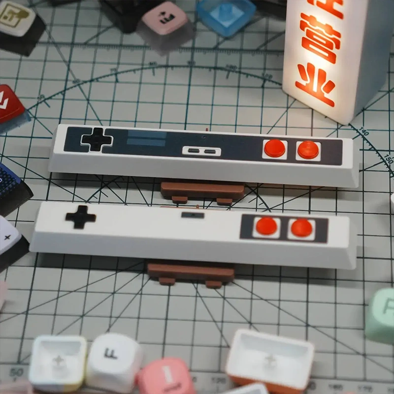 Two Retro Gaming Controller Style Spacebar Keycaps with red buttons and D-pad design on grid mat.