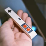 White retro gaming controller spacebar keycap in hand showing D-pad and orange AB buttons, classic 8-bit design