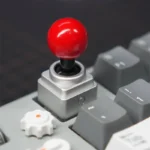 Red-topped metal joystick artisan keycap installed on mechanical keyboard, featuring silver CNC aluminum base.