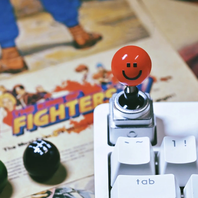 Retro Joystick Metal Artisan Keycap Red joystick ball with black smiley face decal on white keyboard, Street Fighter magazine, playful gaming setup