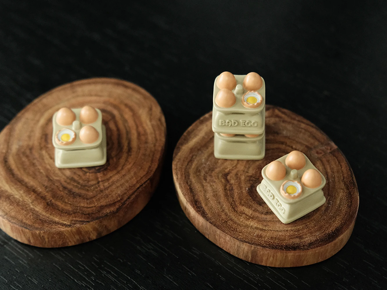 Three egg tray artisan keycaps displayed on wooden coasters with hand-painted miniature eggs and Bad Egg embossed label bases
