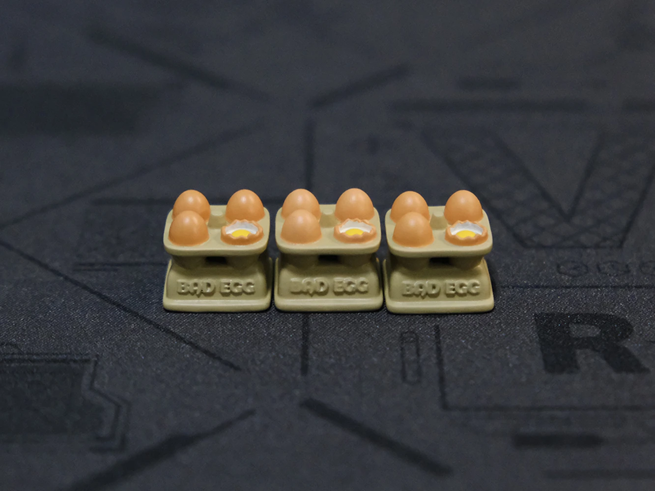 Three egg tray keycaps aligned in row on dark surface displaying Bad Egg labels and hand-painted eggs with yolk details