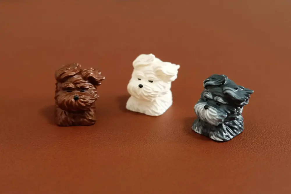 Trio of resin puppy keycaps in brown, white, and black colors on a smooth surface.