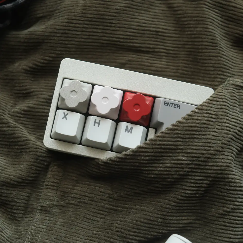 Macro pad with three flower-shaped metal artisan keycaps peeking out of a green corduroy pocket.