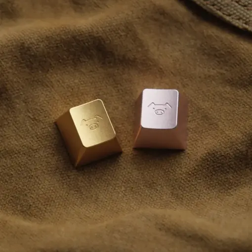 Cute Pig Engraved Metal Artisan Keycaps Gold and Pink Champagne gold and blush pink metal artisan keycaps with a cute pig engraving on a brown fabric background.