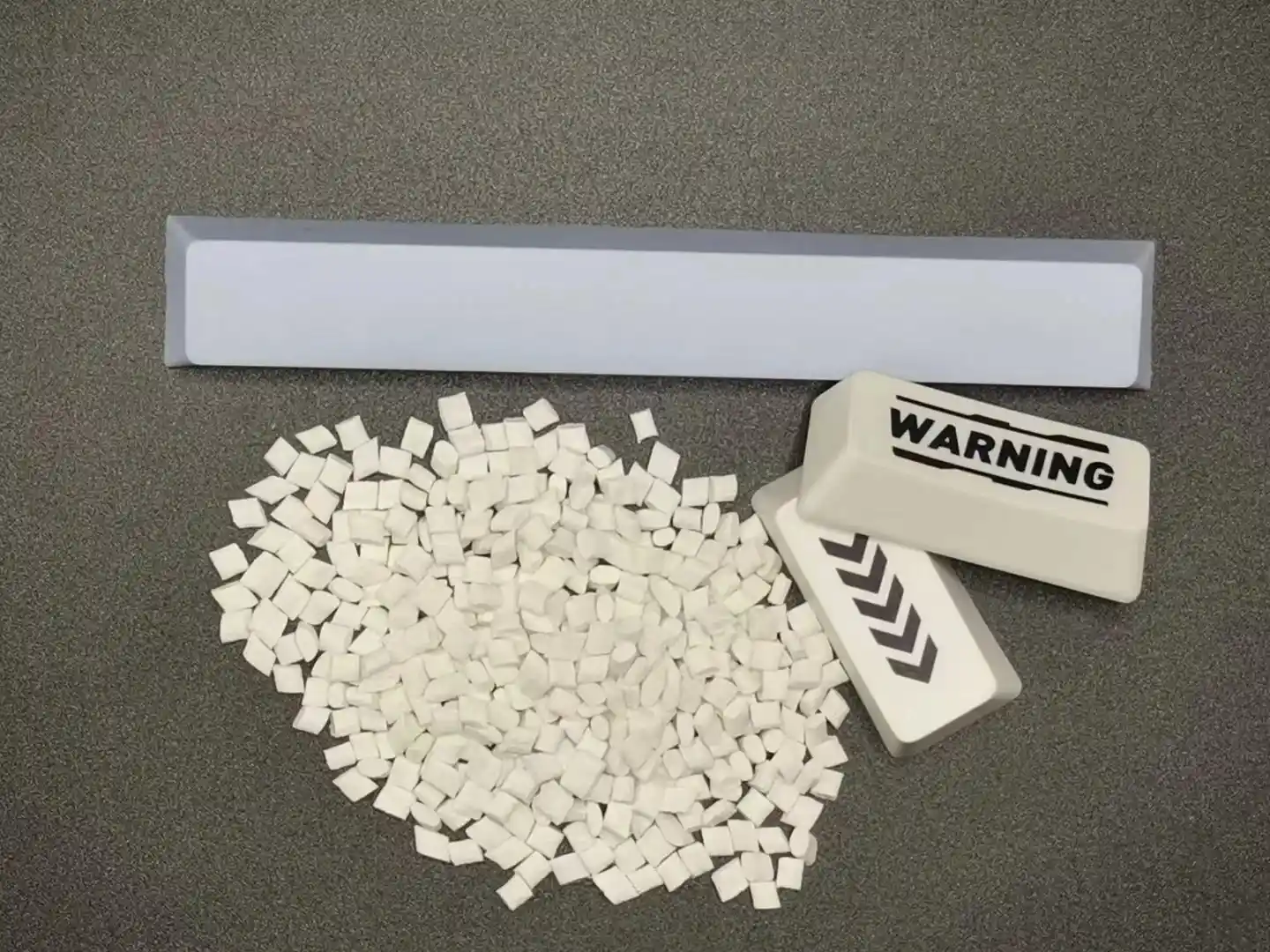 White PBT resin pellets displayed next to finished PBT keycaps and a spacebar, showing the raw material used in manufacturing durable mechanical keyboard keycaps.