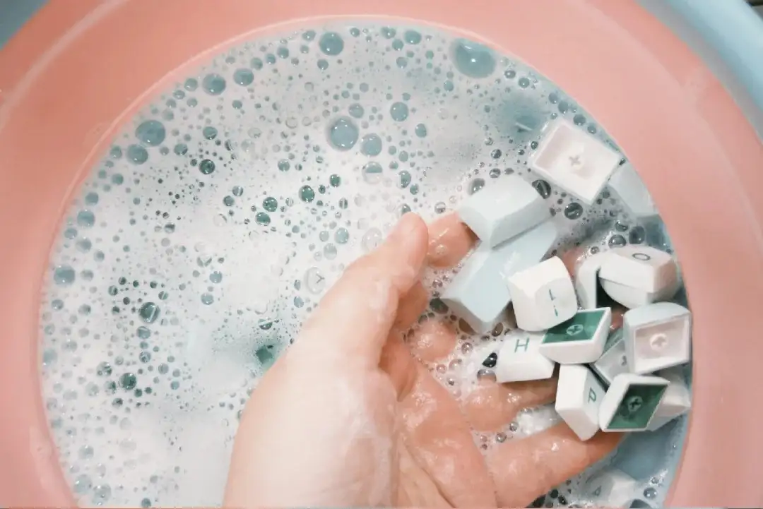 A set of PBT keycaps soaking in a bowl of warm water with mild dish soap, with a wire keycap puller visible beside the bowl