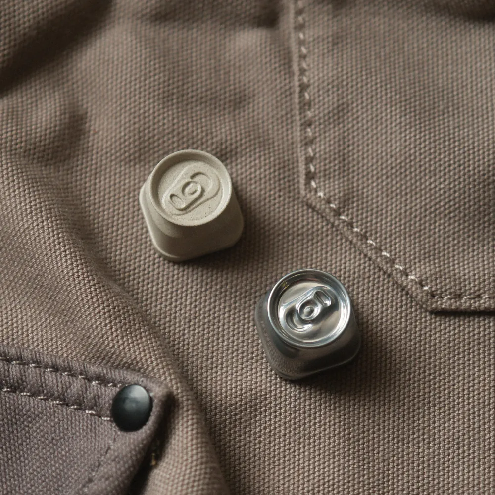 Two metal keycaps for soda cans, one in bright silver and the other in linen color, are displayed on a brown-textured surface.
