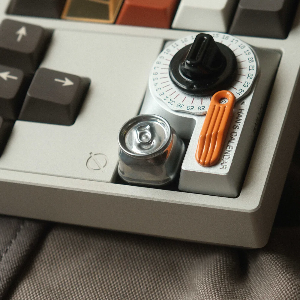 Miniature metal soda can custom ESC keycap in silver finish sitting on a keyboard next to a calendar dial.