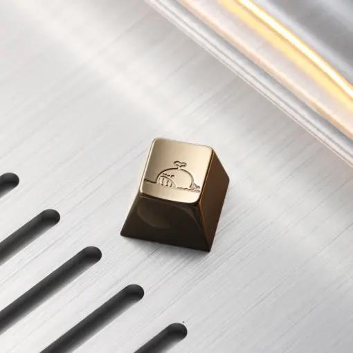 Cute Whale Brass Keycap Close Up Cute whale mirror polished brass artisan keycap resting on a slotted brushed metal background.