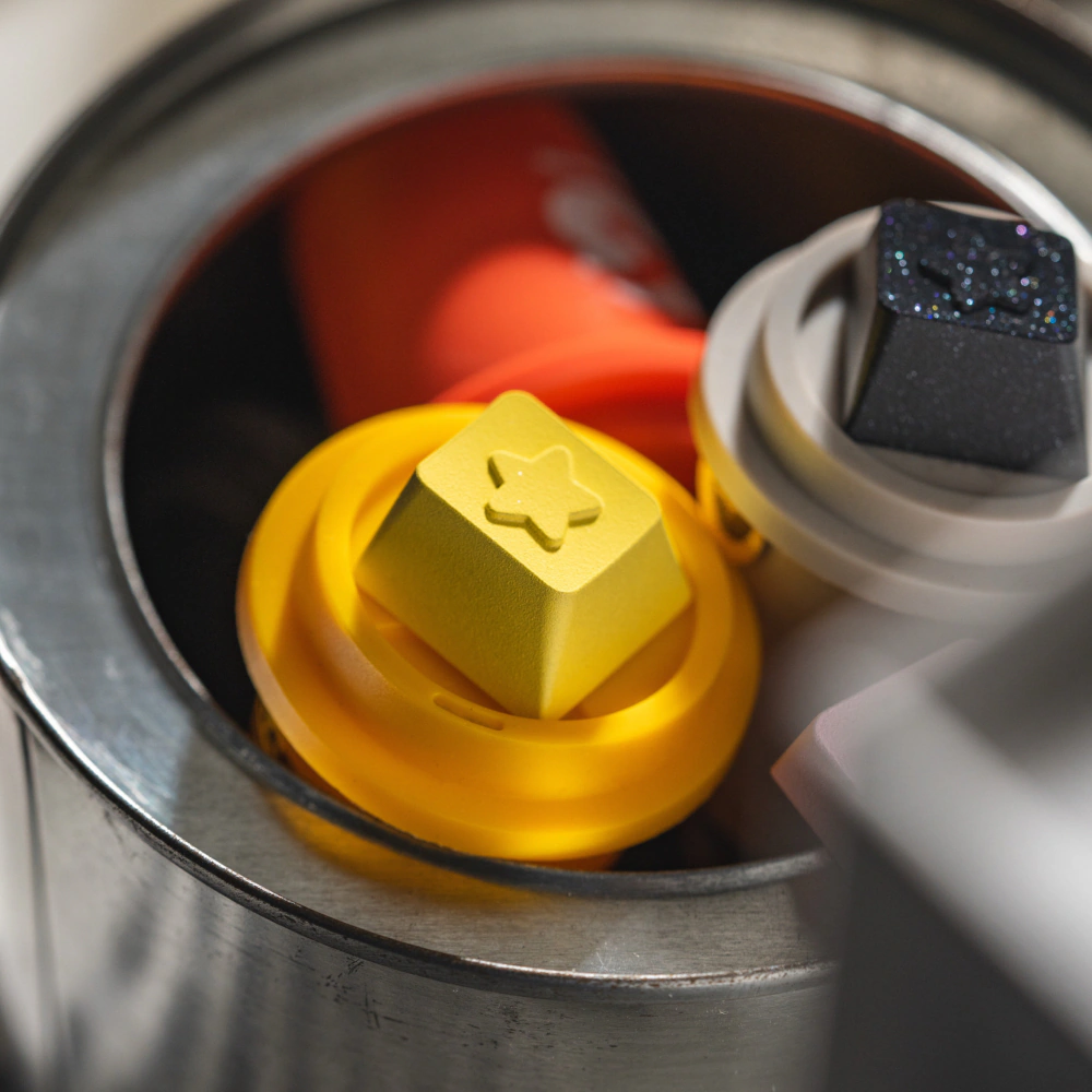 Yellow star themed metal artisan keycap shown close up next to a black keycap.