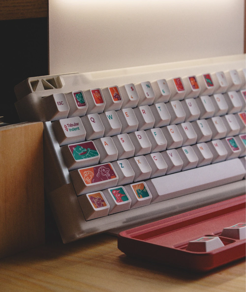 Left side of a white keyboard with colorful creature keycaps and a red tray in front.