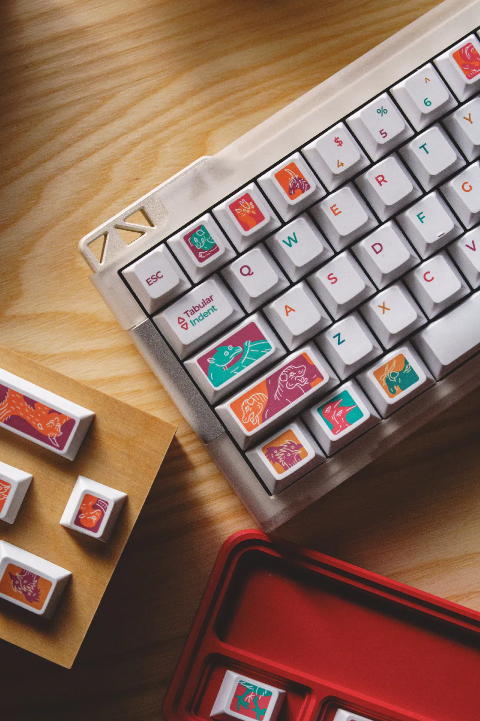 Left side of a white keyboard with colorful creature keycaps, loose keys, and a red tray.
