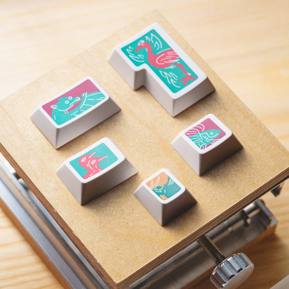 Five white keycaps with colorful creature illustrations displayed on a wooden platform.
