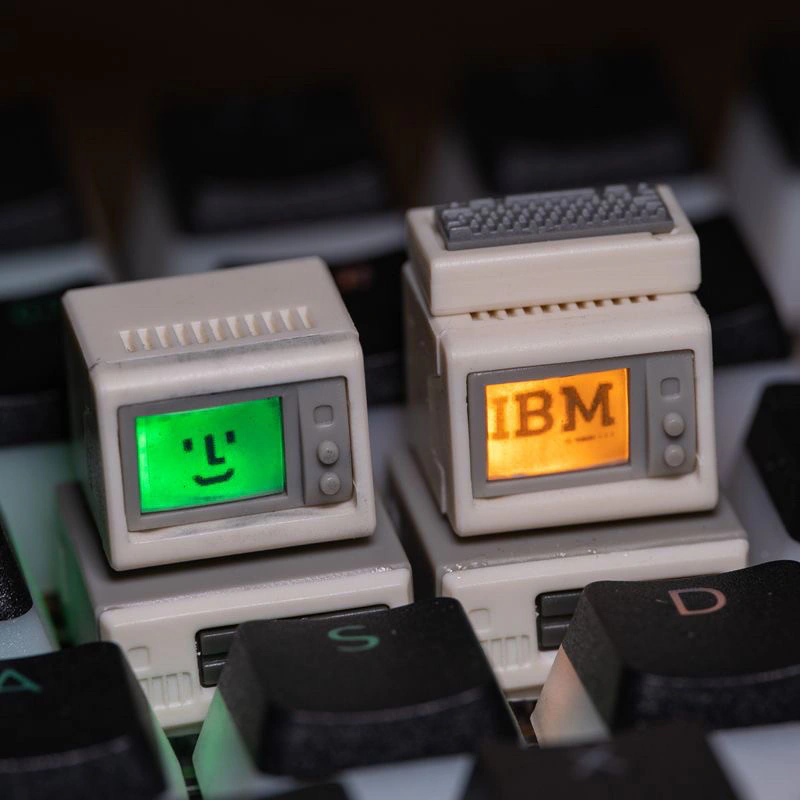 Two beige computer-shaped keycaps on dark keys; the left screen shows a green face and the right shows orange IBM lettering.