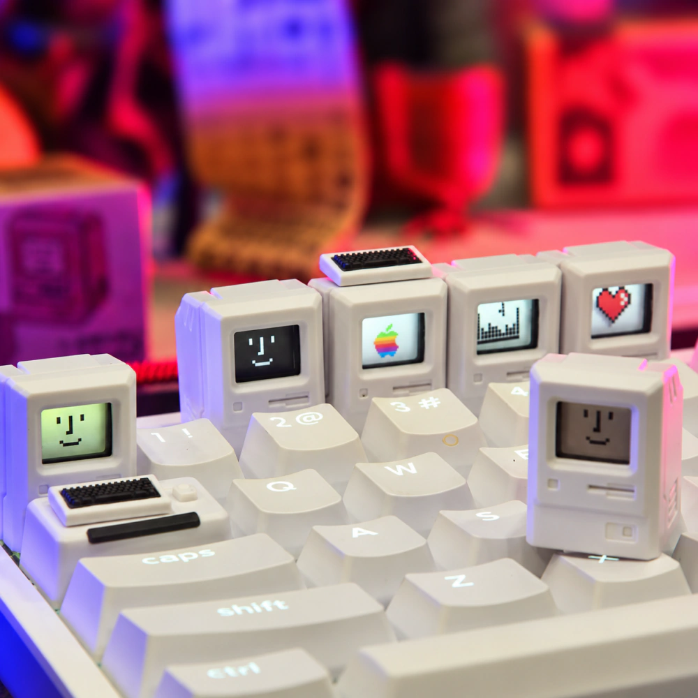 White Macintosh-style keycaps on a white keyboard with pixel face, rainbow, skyline, and heart screen designs.