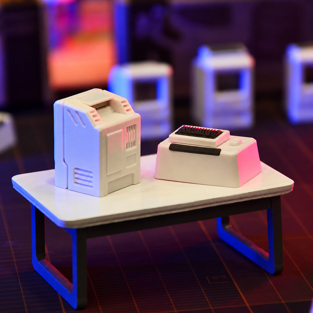 White miniature computer and keyboard pieces on a dark gridded surface, viewed from the side.