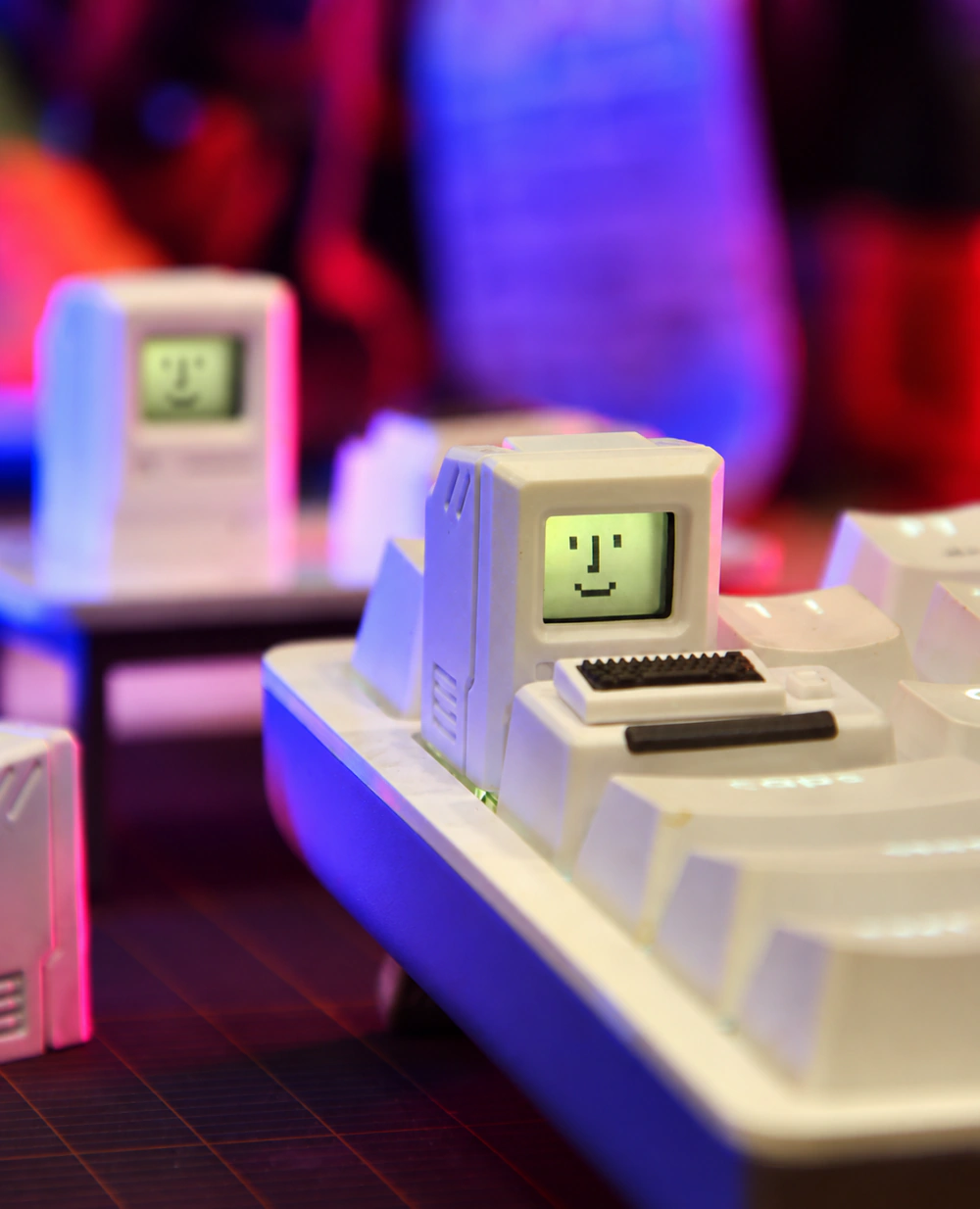 White miniature computer cap on a keyboard, with a pixel face on the screen and the small keyboard piece nearby.